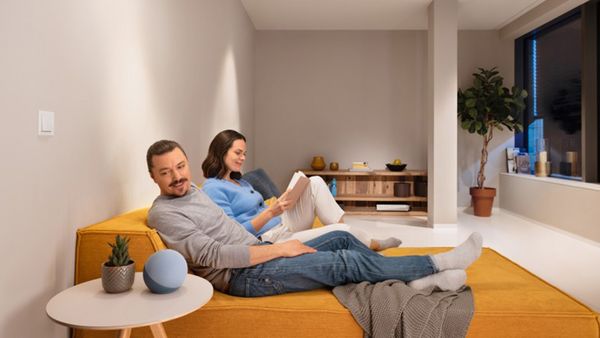 Ein Paar sitzt in einem modernen Wohnraum auf einem gelben Sofa und der Mann interagiert mit einem Alexa Lautsprecher.