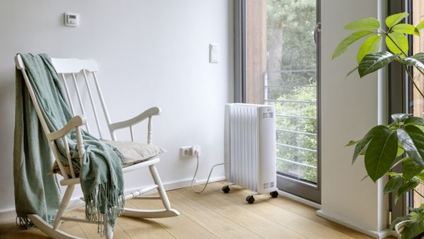 Ein heller Raum mit einem Schaukelstuhl auf dem eine Deckle liegt und einem fahrbarer Elektroradiator steht vor einem Fenster.