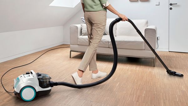 A person uses a Bosch bagless vacuum to clean a small loft space with a couch.