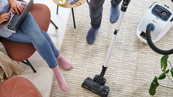 Ein Teppich wird mit einem Bosch Staubsauger gesaugt während eine Frau am Laptop arbeitet.