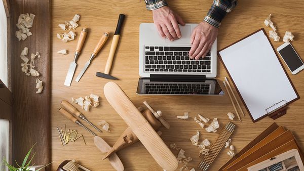 Ein Mann schreibt auf eine Laptop. Dieses steht auf einem Holztisch neben einem Schreibblock mit Handy, sowie Schnitzwerkzeug, Holzspäne und einem Holzflugzeug.