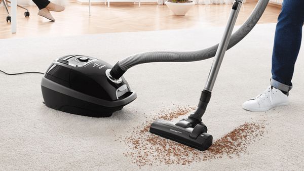 Eine Person saugt mit einem schwarzen Bosch Serie 8 Staubsauger mit Staubbeutel Schmutz von einem hellen Teppichboden.