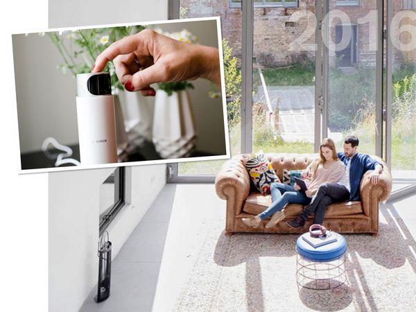 Ein Paar sitzt auf einem braunen Sofa in einem hellen und modernen Wohnzimmer und betrachtet etwas auf einem iPad. Im Vordergrund ist ein Bild in dem eine Hand eine Bosch Überwachungskamera berührt. 