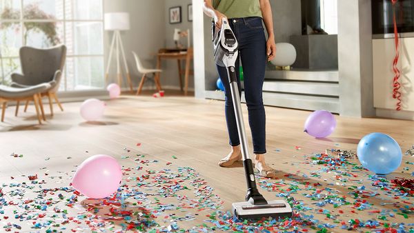 Eine Person saugt mit einem Bosch Akku-Staubsauger den Hartboden eines modernen Wohnraums nach einer Feier. Auf dem Boden liegen bunte Luftballons und Konfetti.