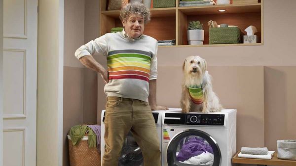 Vīrietis atrodas blakus veļasmašīnai un suns virs veļasmašīnas.