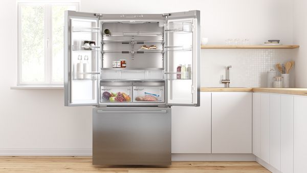 Dans une cuisine blanche et lumineuse, les portes-fenêtres d'un réfrigérateur pose libre sont grandes ouvertes, révélant un intérieur spacieux et bien organisé.