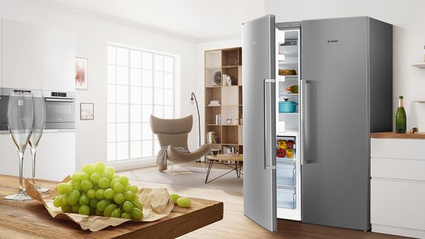 Un grand réfrigérateur-congélateur side-by-side, porte entrouverte, est installé à l'entrée entre une cuisine lumineuse, où des verres à vin et des raisins sont posés sur un comptoir, et l'espace de vie.