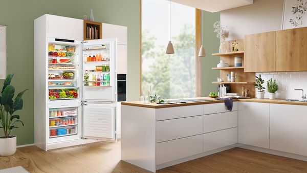Una puerta abierta frente a un mostrador en una cocina amplia y luminosa revela un frigorífico combi XXL incorporado y su abundante contenido.