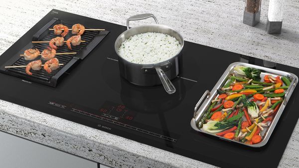 Une table de cuisson à induction dotée d'une plaque teppanyaki pour faire frire des crevettes couvrant deux zones à gauche, une casserole avec du riz au milieu et à droite, une grande plaque sur deux zones où chauffent des légumes.