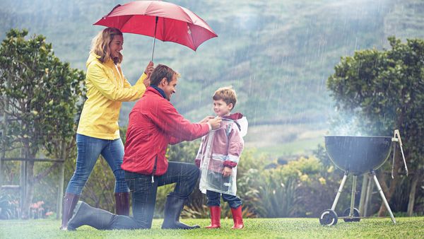 A man adjusts a young child's rain jacket, whilst a woman covers them with an umbrella. A smoking bbq can be seen near to the group. It is clearly raining in the picture.