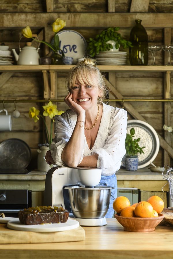 Clodagh McKenna stands at a rustic kichen counter. She leans on a white Bosch CreationLine Stand Mixer. In front of her, there is a cake and a bowl full of oranges.
