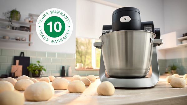 A imagem mostra o robô de cozinha Serie 6 na bancada da cozinha ao lado de várias bolas de massa e do logótipo de garantia de 10 anos sobreposto.