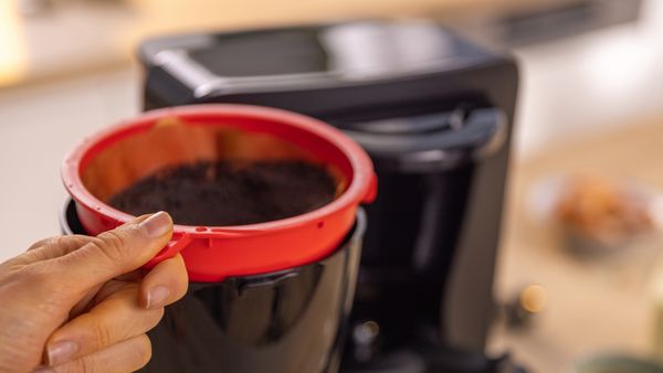 Ръка управлява червен MyMoment въртящ се филтър за кафе с кафе машина на заден план.