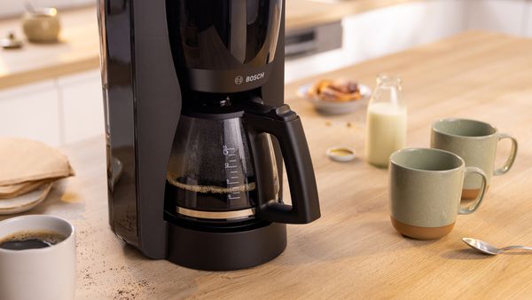 Bosch MyMoment Filterkaffeemaschine mit Glaskanne, die Kaffee enthält, auf einer Küchenarbeitsplatte neben einigen Kaffeetassen.