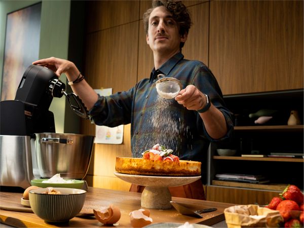 Кухненски робот от Серия 6 върху кухненски плот и човек, който просто поръсва пудра захар върху торта, докато работи с кухненската машина.