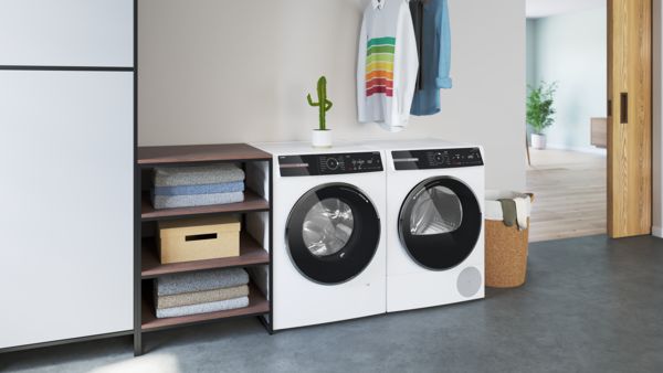 Lavanderia moderna con lavatrice e asciugatrice bianche, scaffale in legno e vestiti appesi, decorata con piante verdi.