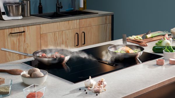 Fresh ingredients are scattered across an island with a built-in induction venting hob where two frying pans of food are cooking.