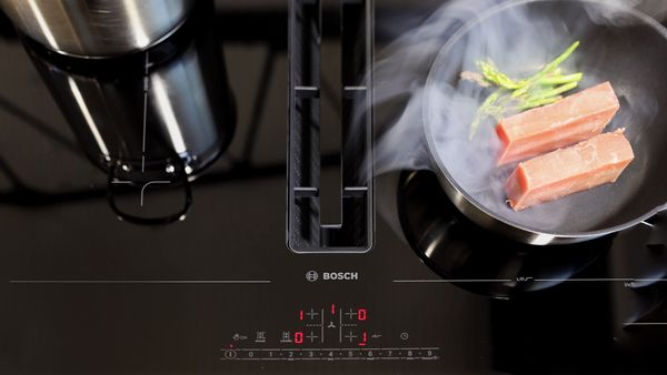 Een close-up van stoom en geurtjes van een koekenpan met zalm die in de ingebouwde afzuigkap van een kookplaat wordt gezogen.
