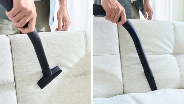 Primo piano di una mano che impugna l'accessorio per interstizi per pulire gli angoli del divano a sinistra. La spazzola per mobili viene usata per aspirare il rivestimento del divano a destra.