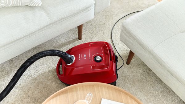 Czerwony, kompaktowy odkurzacz cylindryczny łatwo przesuwa się w wąskiej przestrzeni między dwiema kanapami, aby odkurzyć dywan.
