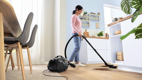 Una mujer utiliza una aspiradora con cable para diferentes tipos de suelos y en una luminosa zona de salón y cocina.