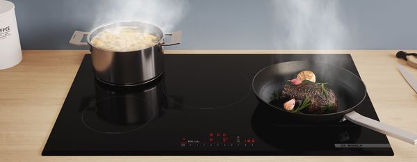 An Bosch induction hob with a pot of asparagus and steak in a pan.
