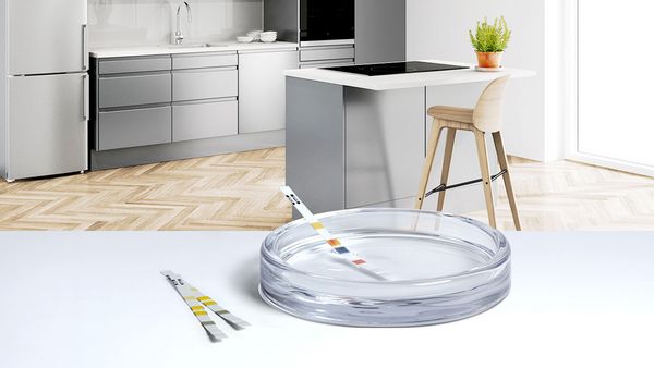 Стъклена купа и тестови ленти за твърдост на водата стоят върху бяла маса със светла, модерна кухня на заден план.
