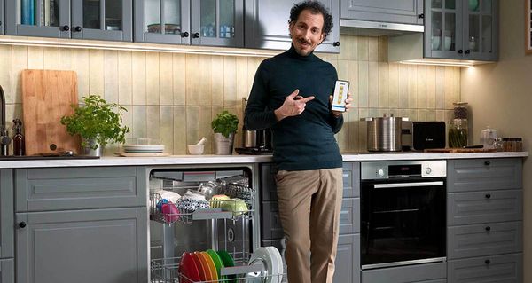 Ein Mann steht in einer Küche neben einem voll beladenen Geschirrspüler und hält ein Smartphone in der Hand.