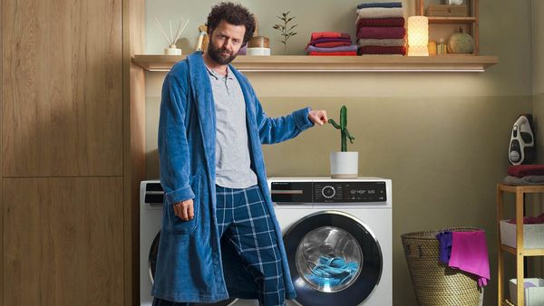 Ein Mann steht in einer Wachküche vor einer Waschmaschine und macht einen Fist Bump oder  Fausstoß mit einem Kaktus, der auf der Waschmaschine steht.