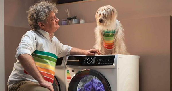 Ein Mann kniet in einer Waschküche vor einem Bosch Wärmepumpentrockner. Er lächelt, weil der energieeffiziente Wärmepumpentrockner Energie spart - gemäß dem EU-Energielabel der höchsten Effizienzklasse, das auch auf seinem Pullover abgebildet ist. Ein Hund im gleichen Pullover sitzt auf dem Wärmepumpentrockner.
