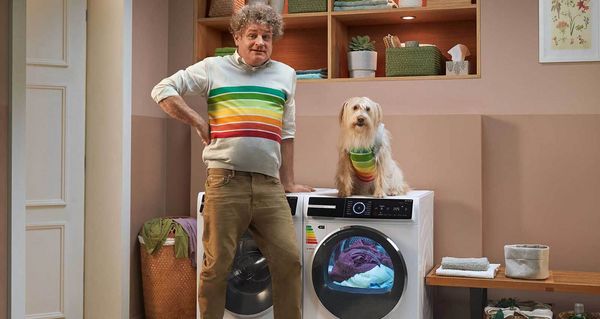 Ein Mann steht in einer Waschküche mit einem Bosch Wärmepumpentrockner. Er lächelt, weil der energieeffiziente Wärmepumpentrockner Energie spart - gemäß dem EU-Energielabel der höchsten Effizienzklasse, das auch auf seinem Pullover abgebildet ist. Ein Hund im gleichen Pullover sitzt auf dem Wärmepumpentrockner.