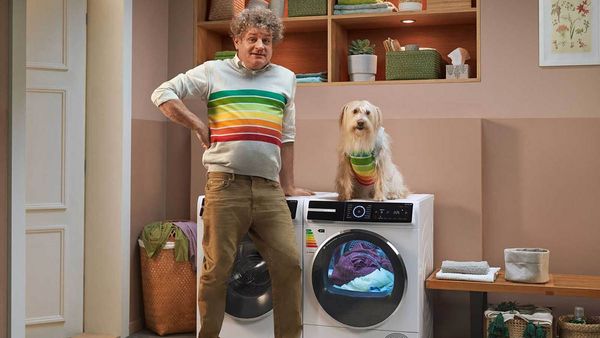Ein Mann steht in einer Waschküche mit einem Bosch Wärmepumpentrockner. Er lächelt, weil der energieeffiziente Wärmepumpentrockner Energie spart - gemäß dem EU-Energielabel der höchsten Effizienzklasse, das auch auf seinem Pullover abgebildet ist. Ein Hund im gleichen Pullover sitzt auf dem Wärmepumpentrockner.