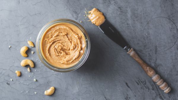 Cashew nut butter in glass jar next to cashew nuts and a knife.