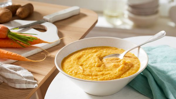Piure de morcovi cu carne de pui într-un bol din ceramică cu lingură, aşezat lângă o planşetă de tăiat pe care se află morcovi.