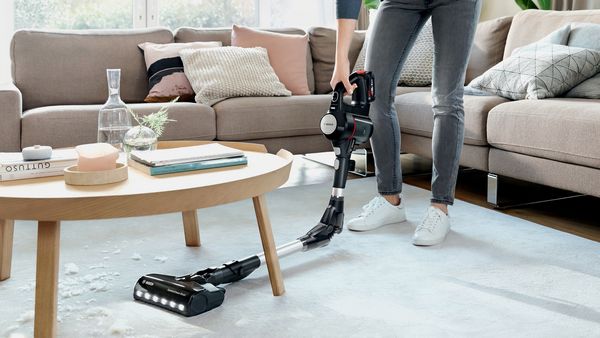 A woman slides a stick vacuum under a coffee table, showing how the tube bends 90 degrees.
