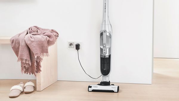 A 2in1 upright vacuum stands charging at an wall socket next to a bench with a scarf and slippers.