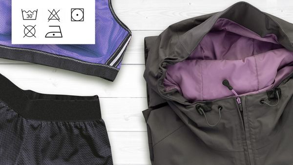 Eine graue Jacke, ein violetter Sport-BH und schwarze Shorts aus atmungsaktivem Gewebe, auf weißen Brettern drapiert.