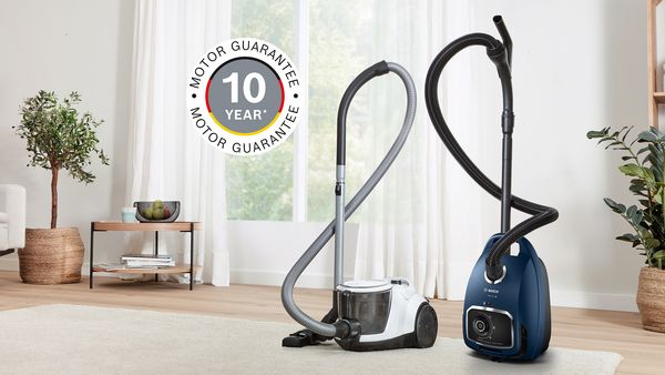 Two Bosch cylinder vacuums bagged and bagless stand on a cream rug in a light, bright living room, next to them the sign for 10-year motor warranty.