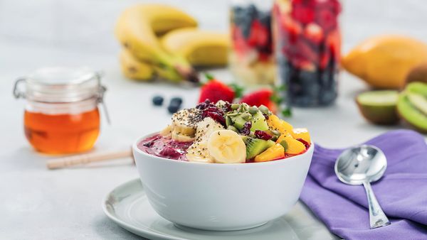 Ceramic bowl filled with blended fruit topped with pieces of fruit in a bowl next to a spoon, purple napkin and jar of honey.