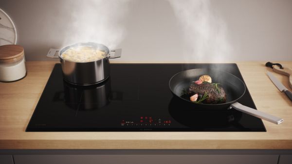 Een inductiekookplaat met aan de linkerkant een grote pan met pasta en de rechterkant een koekenpan met steak, perfect afgestemd op de kookzones.