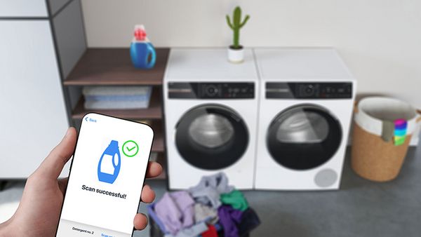 Eine Hand hält ein Smartphone, das intelligente Steuerungen für zwei Waschmaschinen anzeigt, die sich im Hintergrund des Bildes befinden.