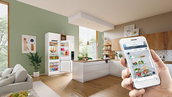 Pametni telefon, ki prikazuje vsebino hladilnika/zamrzovalnika z aplikacijo Home Connect. V ozadju je v kuhinji viden odprt hladilnik/zamrzovalnik, ki ponuja vpogled v priročno in sodobno upravljanje hrane.
