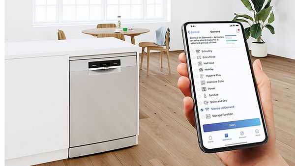 Um ecrã de smartphone apresenta a seleção de modo na aplicação Home Connect, tendo como pano de fundo uma máquina de lavar loiça, uma mesa de jantar e uma ilha de cozinha elegante.