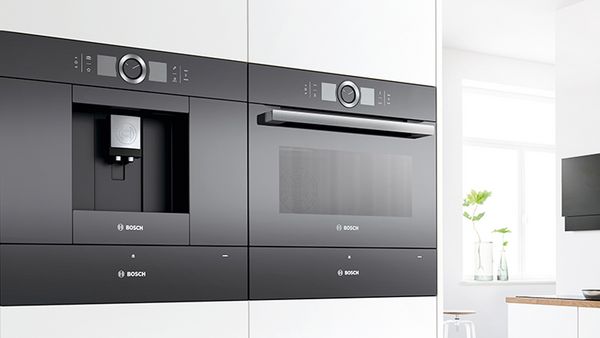 Uma elegante máquina de café preta da Bosch perfeitamente integrada ao lado de um forno moderno, com armários brancos e brilhantes em volta.