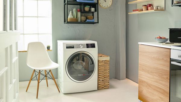 Een wasmachine aan de zijkant van een keuken, met links daarvan een witte stoel. In de rechterhoek zien we een oven en een werkblad met een plant.