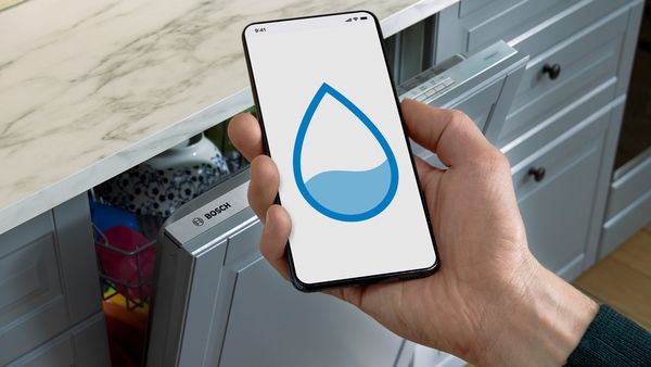 Ръка, която държи мобилен телефон със символ на вода пред кухненски плот с отворена съдомиялна машина.