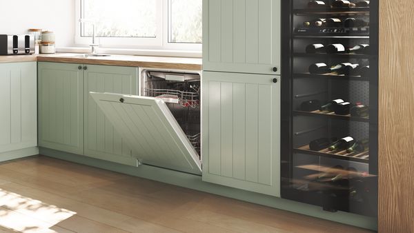 Cocina moderna con armarios de color verde menta, un lavavajillas abierto y un enfriador de vino lleno de botellas junto a una ventana iluminada por el sol.