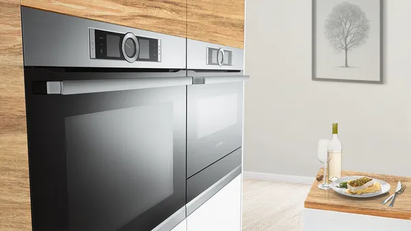 A modern kitchen with a built-in oven and a plate of food, cutlery, bottle of white wine and a glass off to the side on a wooden counter.