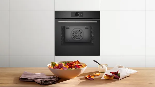 A bowl of colourful vegetable crisps stands on a wooden counter with a built-in oven in the background.