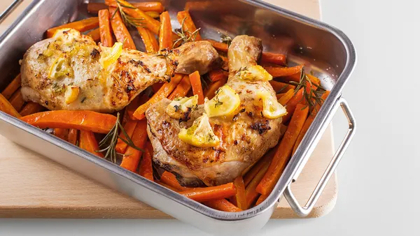 A silver grill pan with cooked chicken pieces on a bed of chopped carrots stands on a wooden pot stand.